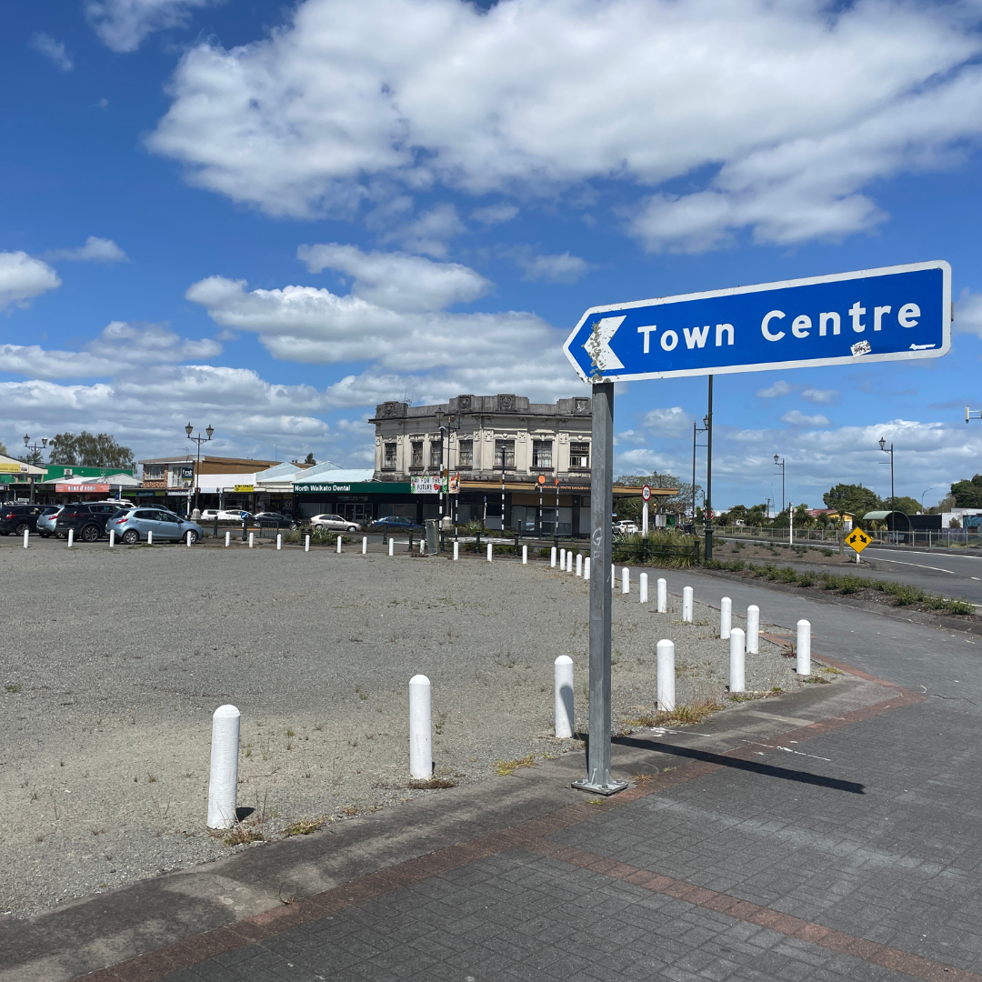 A blue "Town Centre" sign points left above a gray, empty lot with white posts. Behind, buildings line the street, and the sky is bright with fluffy clouds.